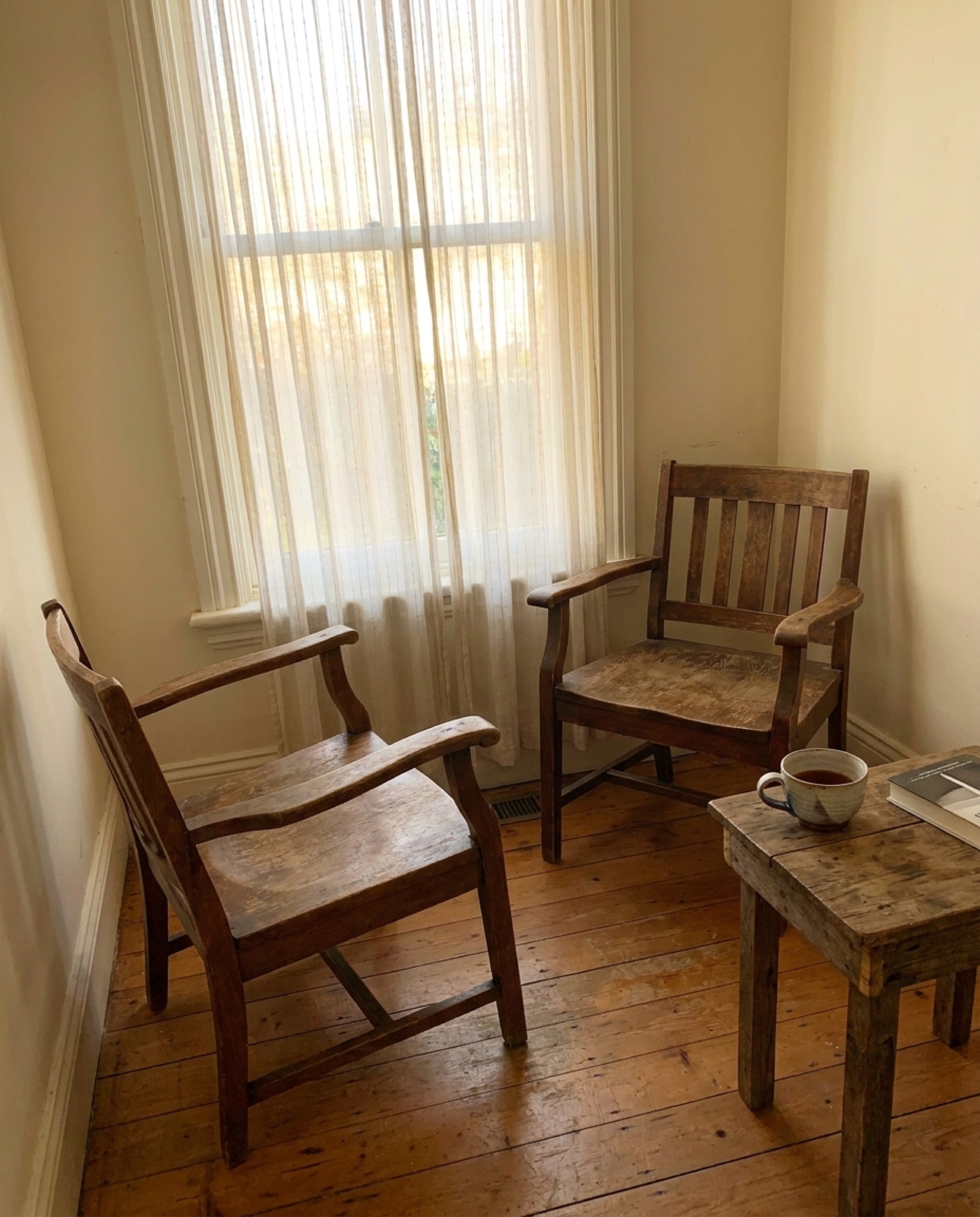 Two empty wooden chairs facing each other in soft afternoon light, a small side table between them holding a single ceramic teacup.